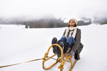 Laughing senior woman sitting on sledge in snow-covered landscape - HAPF02251