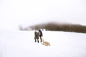 Back view of senior couple with sledge walking side by side in snow-covered landscape - HAPF02253