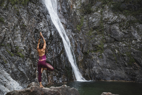 Italien, Lecco, Frau in Baum-Yoga-Pose auf einem Felsen in der Nähe eines Wasserfalls, lizenzfreies Stockfoto