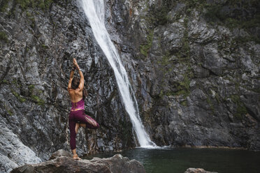 Italy, Lecco, woman doing Tree Yoga Pose on a rock near a waterfall - MRAF00250