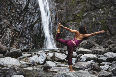 Italien, Lecco, Frau macht Lord of the Dance Yoga Pose auf einem Felsen in der Nähe eines Wasserfalls, lizenzfreies Stockfoto