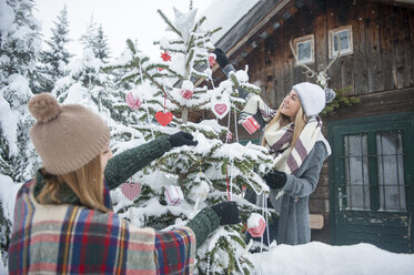 Österreich, Altenmarkt-Zauchensee, zwei junge Frauen schmücken Weihnachtsbaum an Holzhaus - HHF05500