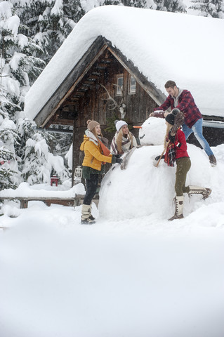 Österreich, Altenmarkt-Zauchensee, Freunde bauen einen großen Schneemann am Holzhaus, lizenzfreies Stockfoto