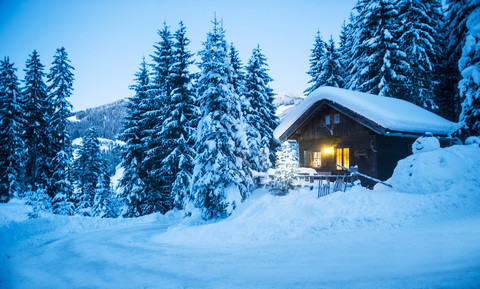 Österreich, Altenmarkt-Zauchensee, Schlitten, Schneemann und Weihnachtsbaum am beleuchteten Holzhaus im Schnee in der Abenddämmerung, lizenzfreies Stockfoto