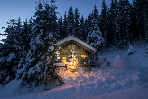 Österreich, Altenmarkt-Zauchensee, Schlitten, Schneemann und Weihnachtsbaum an beleuchtetem Holzhaus im Schnee bei Nacht, lizenzfreies Stockfoto