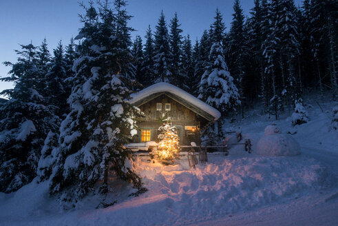 Österreich, Altenmarkt-Zauchensee, Schlitten, Schneemann und Weihnachtsbaum an beleuchtetem Holzhaus im Schnee bei Nacht - HHF05516