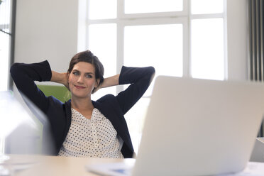 Happy businesswoman sitting in office, looking at laptop with hands behind head - FKF02607