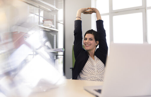 Happy businesswoman sitting in office, looking at laptop, stretching - FKF02609