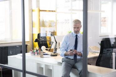 Mature businessman sitting office , using smartphone, reading messages - FKF02613