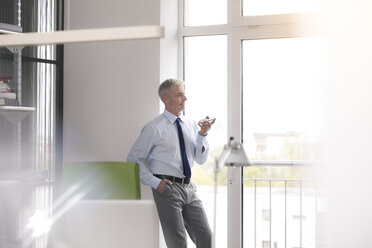 Mature businessman standing in office, using smartphone - FKF02669