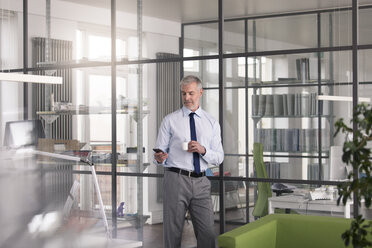 Businessman standing in office, drinking office, using smartphone - FKF02674