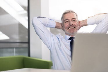 Businessman working in office, looking at laptop with hands behind head - FKF02686