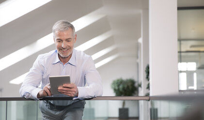 Erfolgreicher Geschäftsmann steht am Geländer in seinem Büro und benutzt ein digitales Tablet - FKF02690