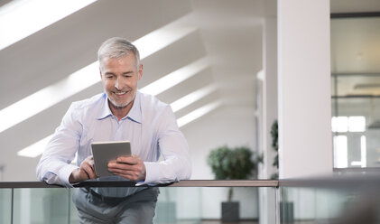 Successful businessman standing at railing in his office, using digital tablet - FKF02690