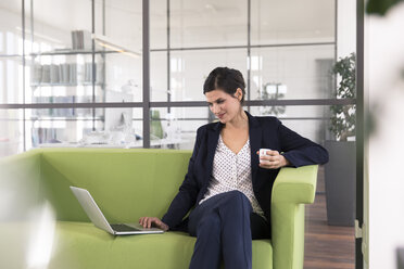 Geschäftsfrau mit Laptop, die auf einer Couch sitzt und im Büro Kaffee trinkt - FKF02699