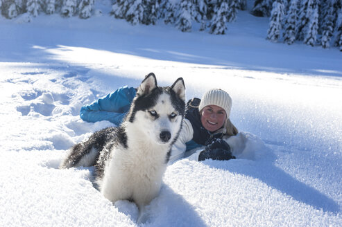 Österreich, Altenmarkt-Zauchensee, Porträt einer lächelnden jungen Frau mit Hund im Schnee liegend - HHF05521