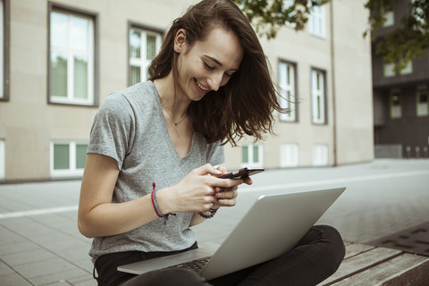 Glückliche junge Frau sitzt auf einer Bank und benutzt Smartphone und Laptop, lizenzfreies Stockfoto