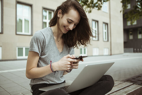 Glückliche junge Frau sitzt auf einer Bank und benutzt Smartphone und Laptop - FEXF00287