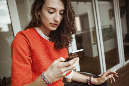 Young woman checking smartphone on balcony smoking a cigarette - FEXF00295