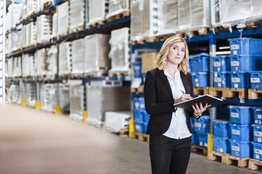 Blond businesswoman standing in storehouse, writing in notebook - DIGF03072