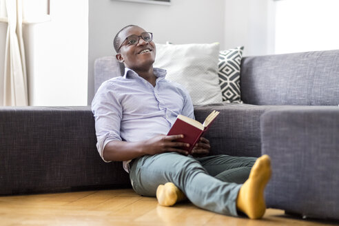 Glücklicher junger Mann sitzt auf dem Boden im Wohnzimmer und liest ein Buch - MMAF00178