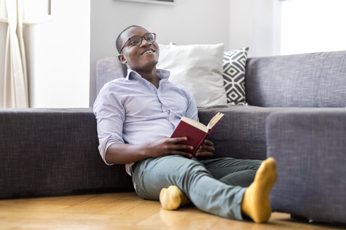 Happy young man sitting on the floor in the living room reading book - MMAF00178