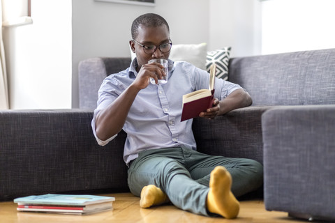 Young man sitting on the floor in the living room reading book and drinking glass of water stock photo