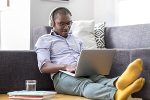 Junger Mann sitzt auf dem Boden im Wohnzimmer mit Laptop und Kopfhörern - MMAF00180
