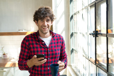 Junger Unternehmer steht in der Firmenküche, trinkt Kaffee, benutzt sein Smartphone - SPCF00223