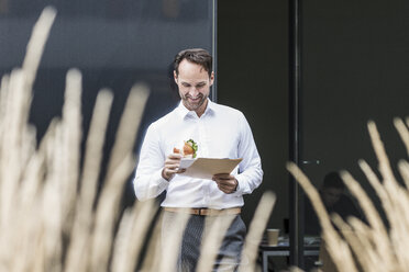 Lächelnder Geschäftsmann mit Sandwich beim Lesen von Dokumenten vor einem Bürogebäude - UUF12069