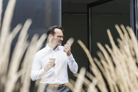 Geschäftsmann mit Kaffee zum Mitnehmen und Mobiltelefon vor einem Bürogebäude, lizenzfreies Stockfoto