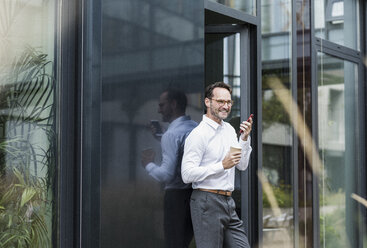 Businessman with coffee to go using cell phone in front of office building - UUF12071