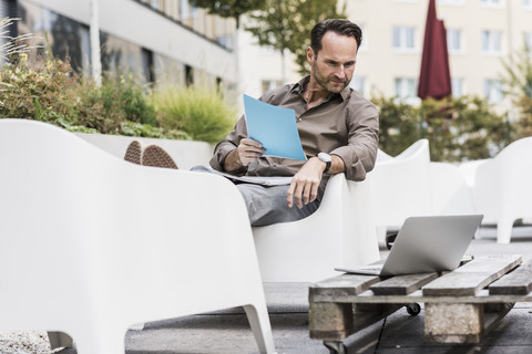 Man with documents sitting on terrace looking at laptop stock photo