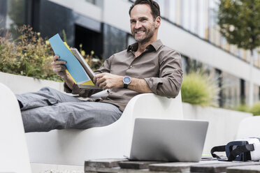 Lächelnder Geschäftsmann mit Dokument, Laptop und Virtual-Reality-Brille auf der Terrasse sitzend - UUF12109