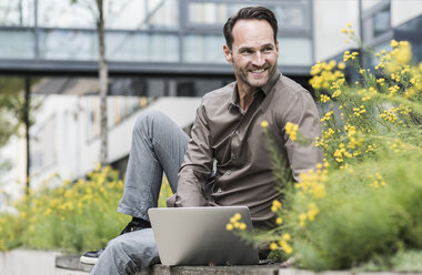 Porträt eines lächelnden Geschäftsmannes bei der Arbeit mit einem Laptop im Freien - UUF12112