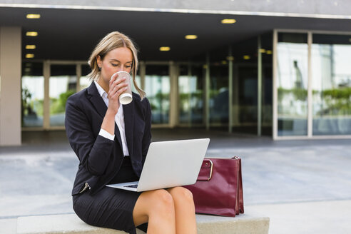 Geschäftsfrau mit modischer Ledertasche und Kaffee zum Mitnehmen sitzt auf einer Bank und schaut auf ihren Laptop - MGIF00184
