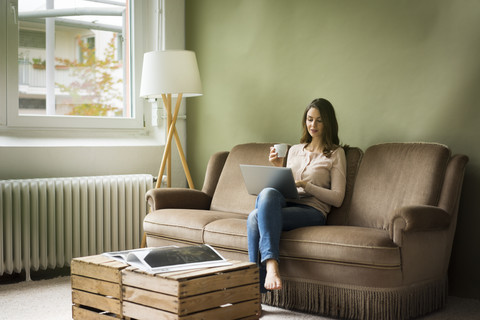 Young woman sitting on couch with cup of coffee using laptop stock photo
