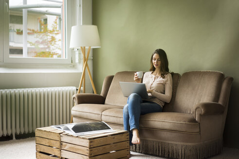 Junge Frau sitzt mit einer Tasse Kaffee auf der Couch und benutzt einen Laptop - MOEF00186