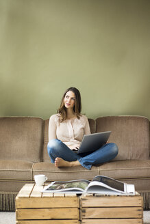 Lächelnde junge Frau sitzt auf einer Couch und benutzt einen Laptop - MOEF00187