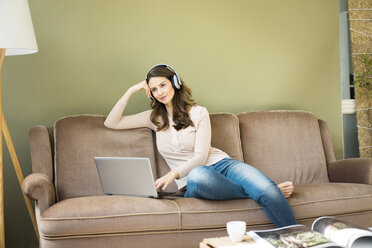 Junge Frau mit Kopfhörern sitzt auf der Couch und benutzt einen Laptop - MOEF00188