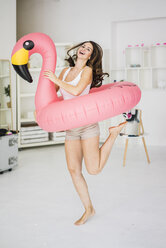 Porträt einer lachenden jungen Frau, die zu Hause mit einem Flamingo-Schwimmspielzeug tanzt - MOEF00210
