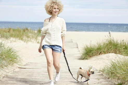 Porträt einer lächelnden jungen Frau, die mit ihrem Hund am Strand Gassi geht - TSFF00142