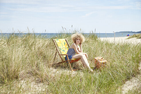 Junge Frau sitzt auf einem Strandkorb in den Dünen und beobachtet etwas - TSFF00149