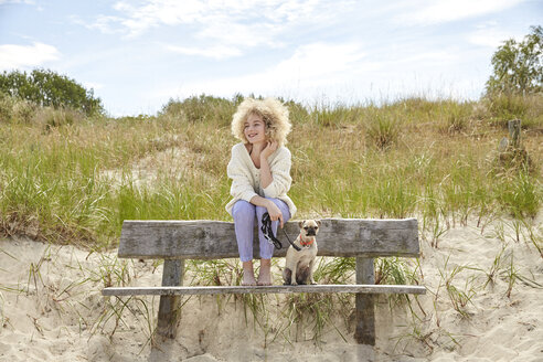 Porträt einer glücklichen jungen Frau, die mit ihrem Hund auf einer Bank in den Dünen sitzt - TSFF00153
