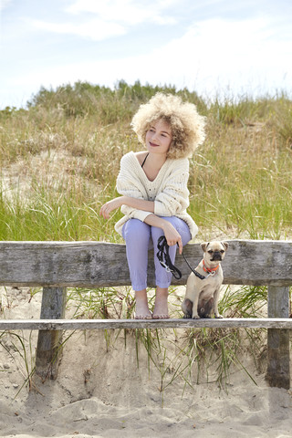 Porträt einer lächelnden jungen Frau, die mit ihrem Hund auf einer Bank in den Dünen sitzt, lizenzfreies Stockfoto