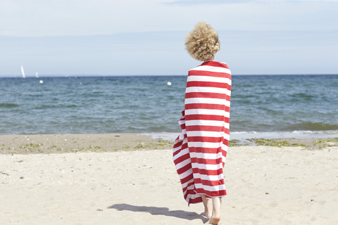 Rückenansicht einer jungen Frau, die in ein Strandtuch eingewickelt vor dem Meer steht, lizenzfreies Stockfoto