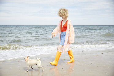 Lächelnde junge Frau in Regenmantel und Gummistiefeln geht mit ihrem Hund am Meer spazieren - TSFF00177