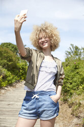 Porträt einer lächelnden jungen blonden Frau, die ein Selfie mit ihrem Smartphone macht - TSFF00194