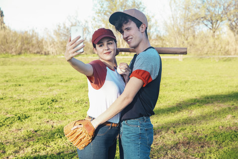 Lächelndes junges Paar mit Baseball-Ausrüstung macht ein Selfie im Park, lizenzfreies Stockfoto