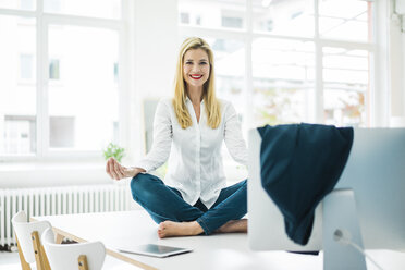 Lächelnde Geschäftsfrau sitzt auf dem Schreibtisch im Büro und übt Yoga - MOEF00228